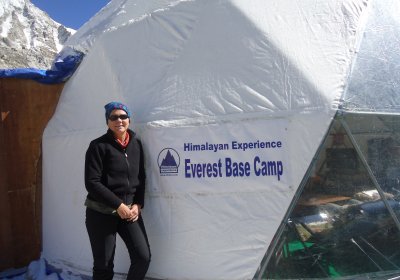 Debra Jackson is stood next to a white dome that says &#039;Everest Base Camp&#039; with mountain in background. Debra is dressed all in black thermals with sunglasses on.