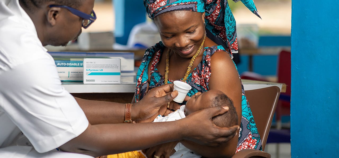 Healthcare worker administering an oral vaccine to an infant held by a caregiver in a clinic setting, with medical supplies and boxes visible in the background. Credit: Stella Abli PSI Cote d'Ivoire