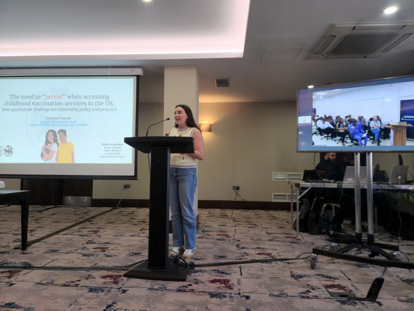 Women standing at lecturn with powerpoint presentation on projector behind her, tv showing remote participants next to her.