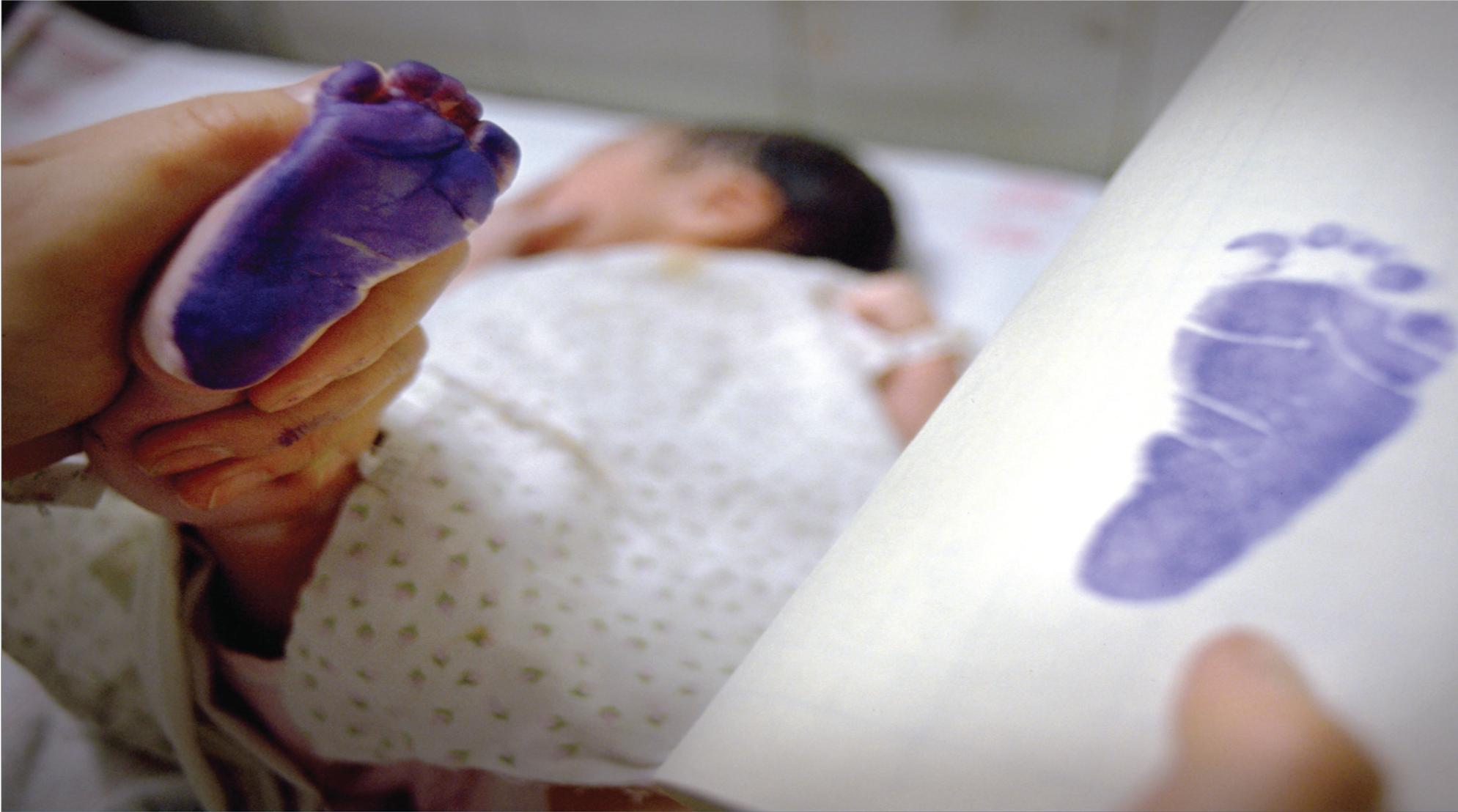 A picture of a newborns footprint being recorded. In foreground is the baby's foot with purple ink on and the footprint on a piece of paper, newborn is blurred in backfround