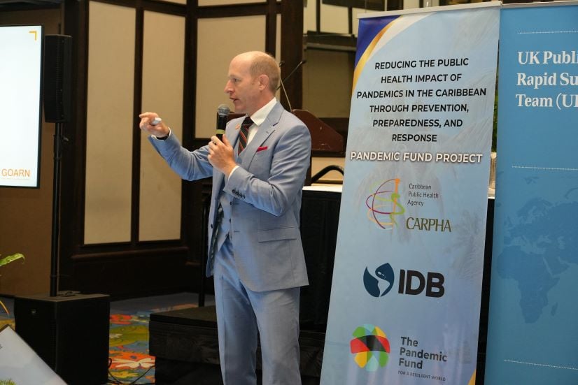 UK-PHRST Ed Newman addressing delegates, standing in front of CARPHA and UK-PHRST baneners.