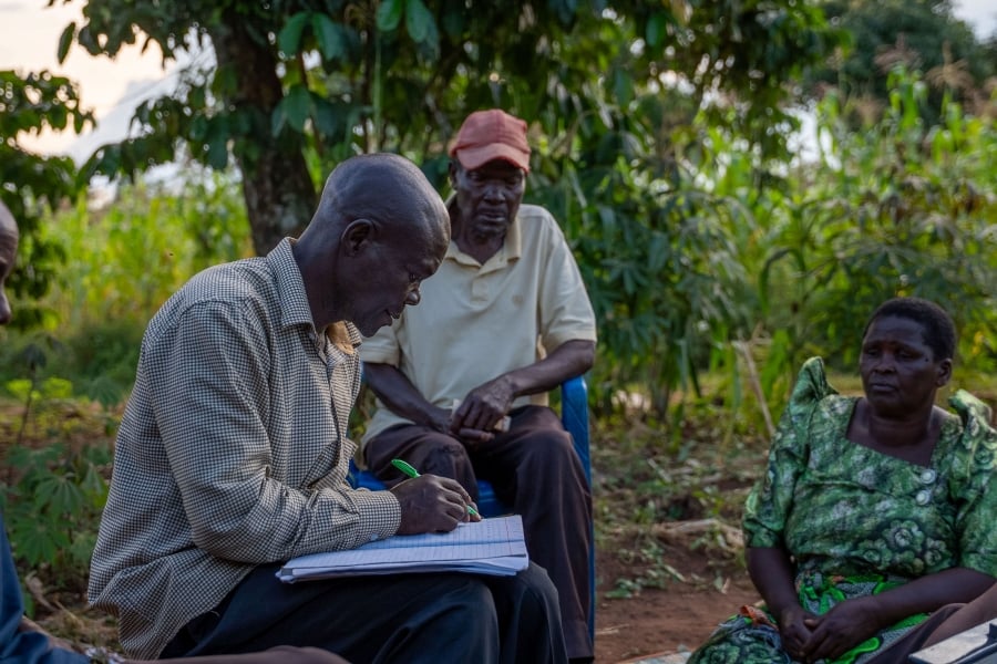 A PLA-D group member in Luuka is writing down notes on a paper at the group meeting. Two other members of the group are sitting next to him and looking on. They are outside, with trees and other foliage around them.