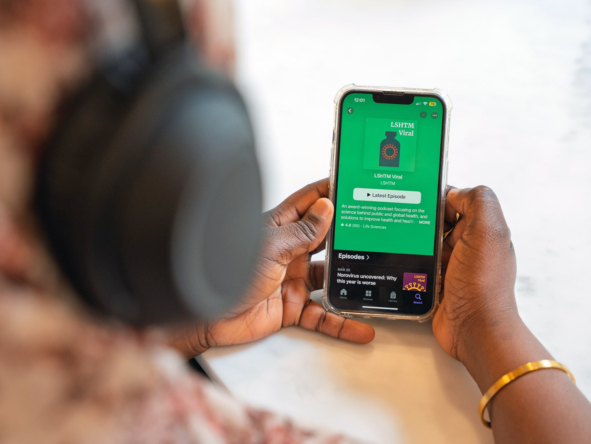 image of a woman facing away from the camera wearing headphones, holding a mobile phone with the podcast 'LSHTM Viral' showing on the screen, in a white room