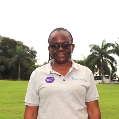 Flavia is wearing glasses and a grey polo t-shirt. She is standing in a grassy lawn and ornamental palm trees are visible behind her.