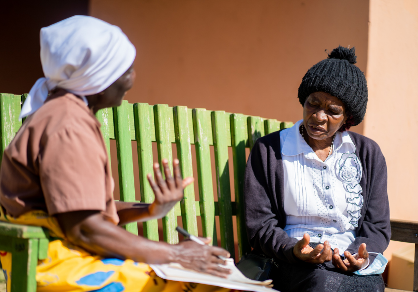 A Friendship Bench Grandmother sitting with her client on simple wooden park bench, using an evidence-based approach to probelm-solving therapy to get people out of depression in Zimbabwe.