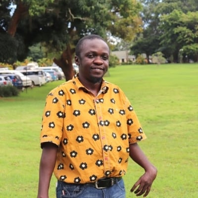 Geofrey is wearing a yellow ochre shirt with black flowers printed on it. He is standing on a grassy lawn with trees in the background. Cars are parked behind the trees.