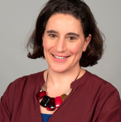 A headshot of Hannah Kuper, who has a short, dark hair and is smiling. She is wearing a statement necklace and maroon top.