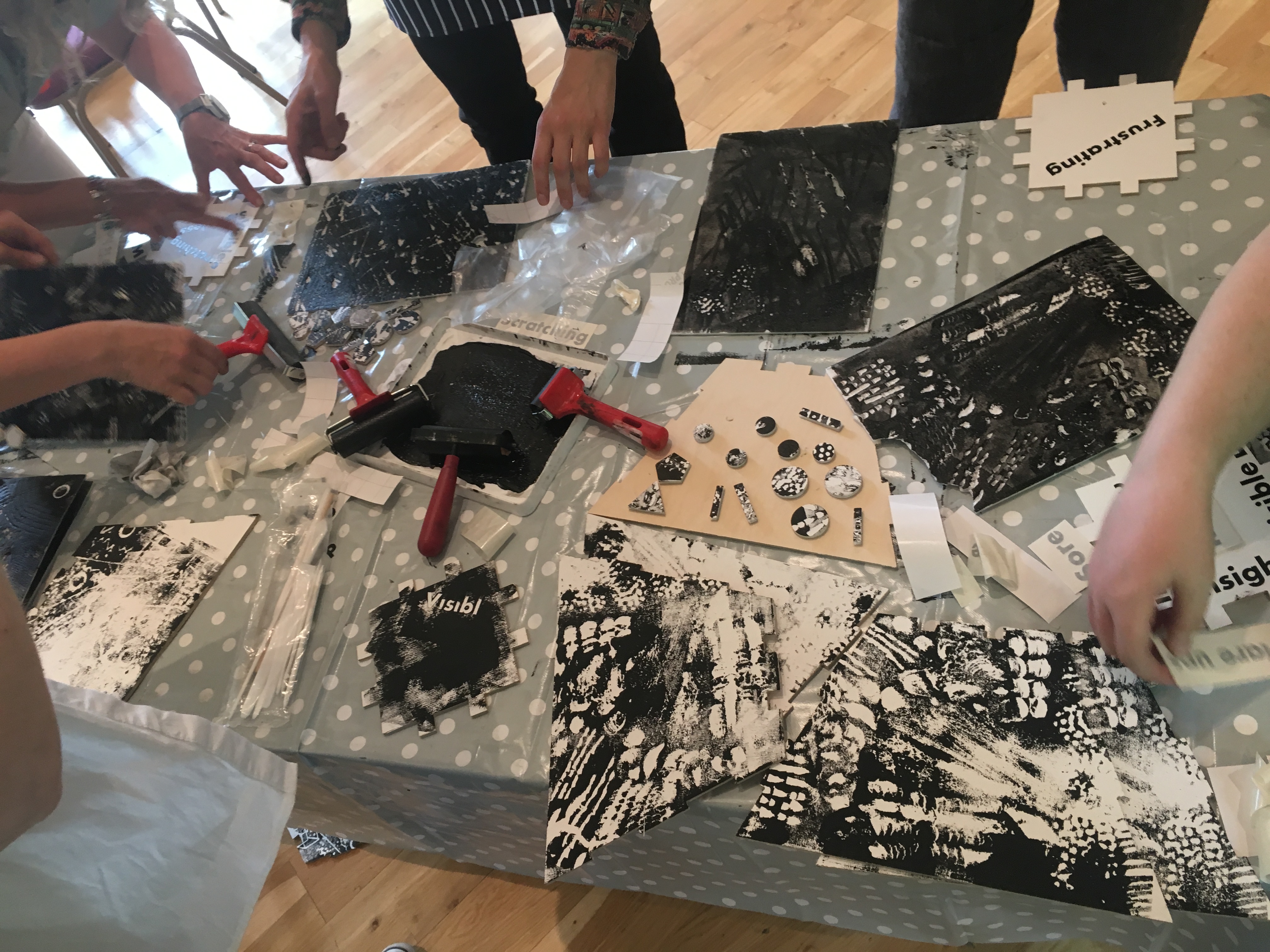Workshop participants are gathered around a table. Their hands are visible and they are printmaking, transferring pattern from Styrofoam on to wooden panels.