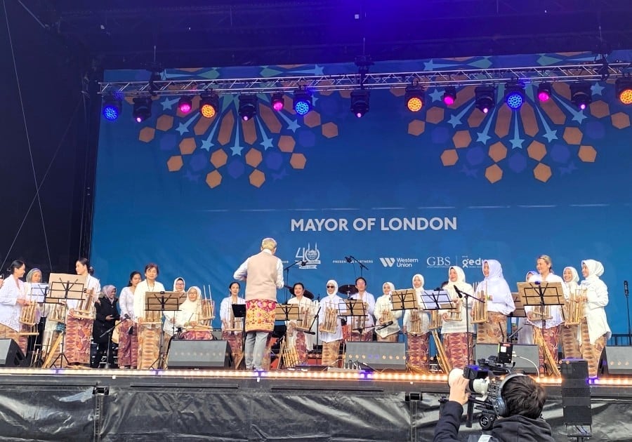 A group of Indonesian women playing in an angklung band at the Eid on the Square celebrations. They are on-stage wearing pale cream outfits with instruments and sheet music stands in front of them.