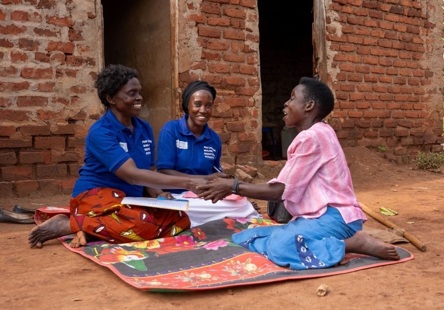 Three women sitting on mat outside a house in Luuka district, Uganda. The two women wearing blue t-shirts are from the Village Health Team and are conducting outreach activities to support people with disabilities.