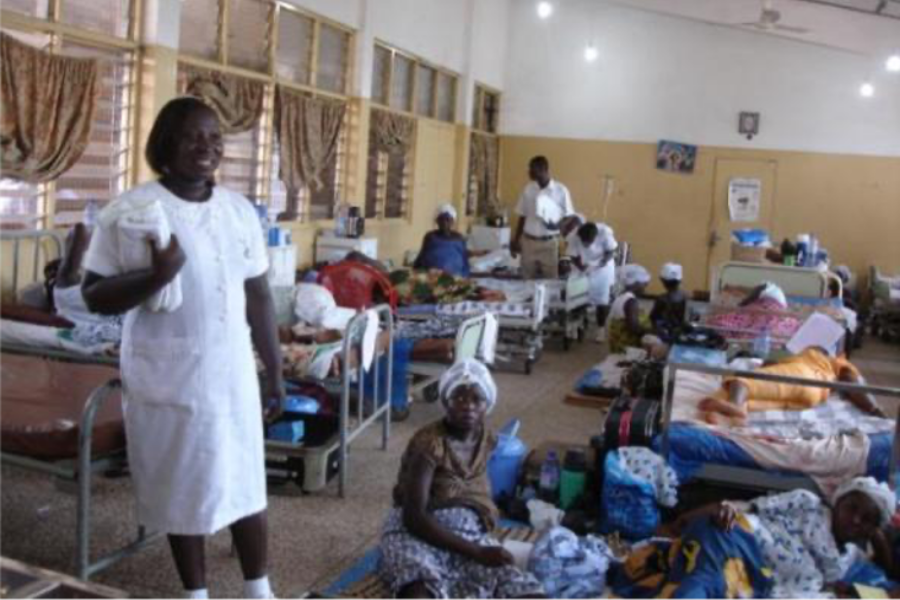 A busy maternity ward in Tanzania. A health care professional dressed in a white dress and smiling is on the left hand side and women are dotted all around sat on beds and on the floor.