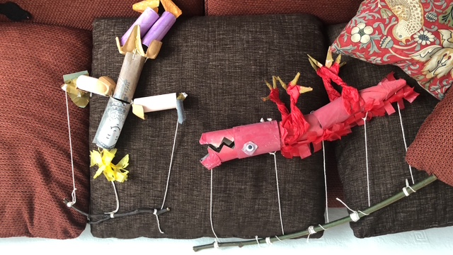 Two puppets, one a red dinosaur and one a silver person made out of carboard sit against a red couch. 