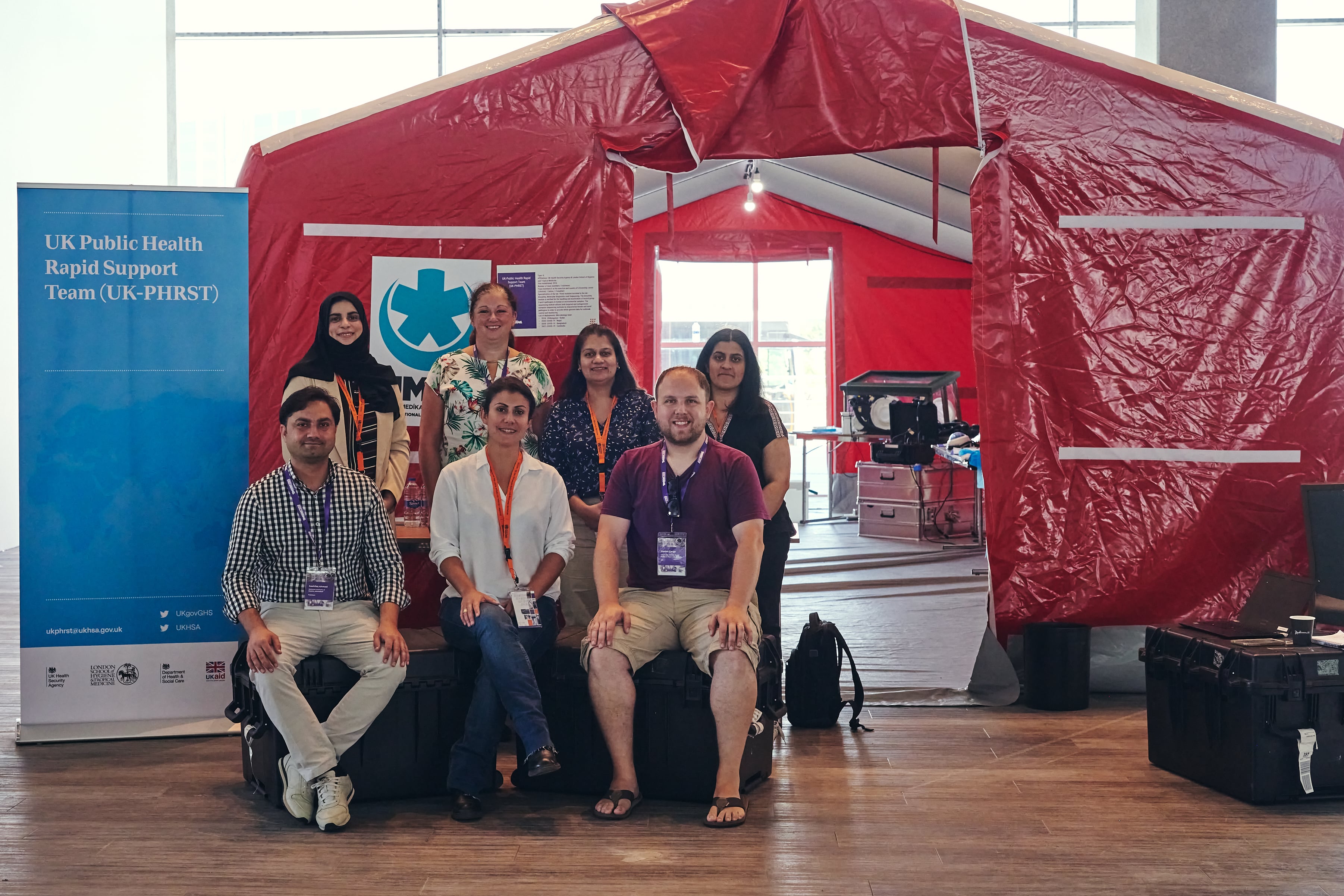 UK-PHRST Microbiology team sitting in front of RRML tent with UK-PHRST banner in the background. 