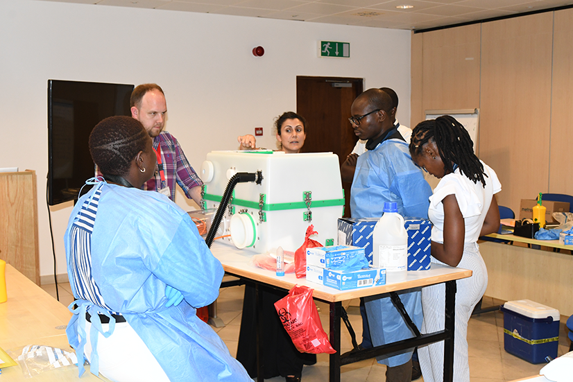 UK-PHRST staff members shows how to use microbiology equipment to Kenya KEMRI Wellcome trust colleagues who are dressed in PPE standing watching demonstration. 