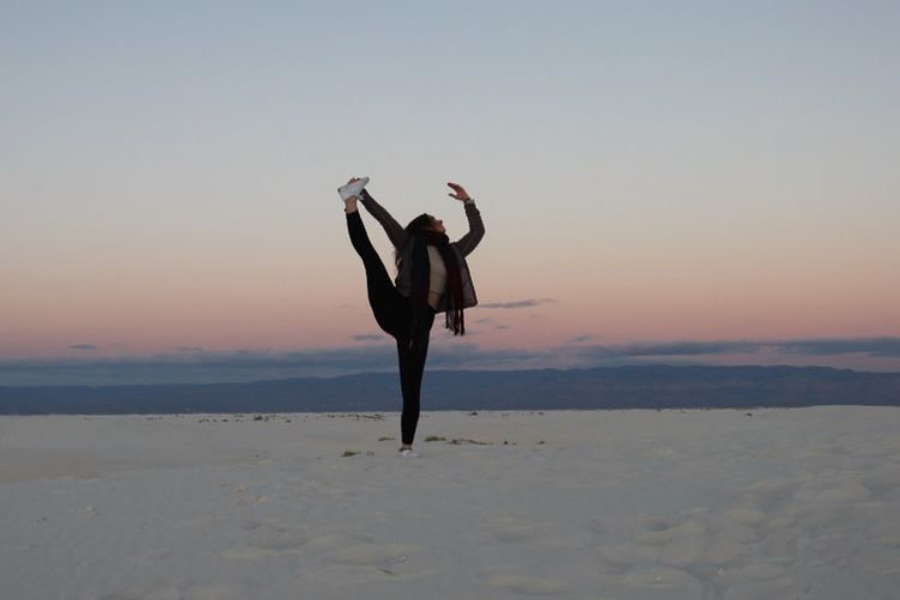 Sabrina is holding one leg up in the air and standing on the other leg on a desert-like landscape at dusk