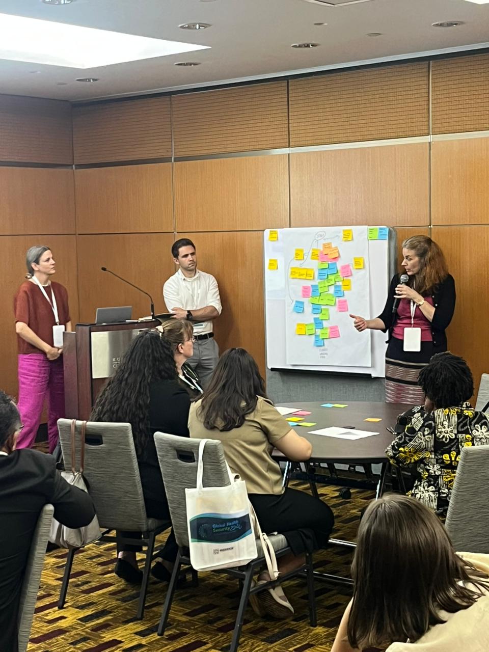 People standing in front of a whiteboard with sticky notes on it, speaking into microphone to conference room. 