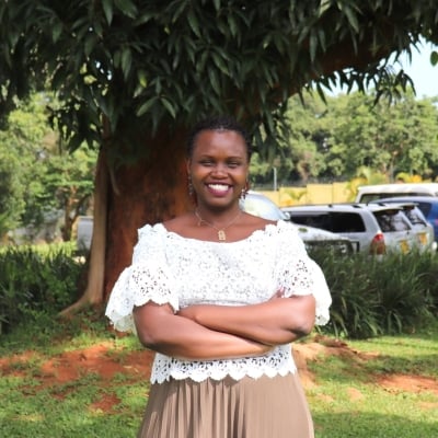 Sylvia is standing with her arms crossed under a tree. She is wearing a white top and beige skirt.