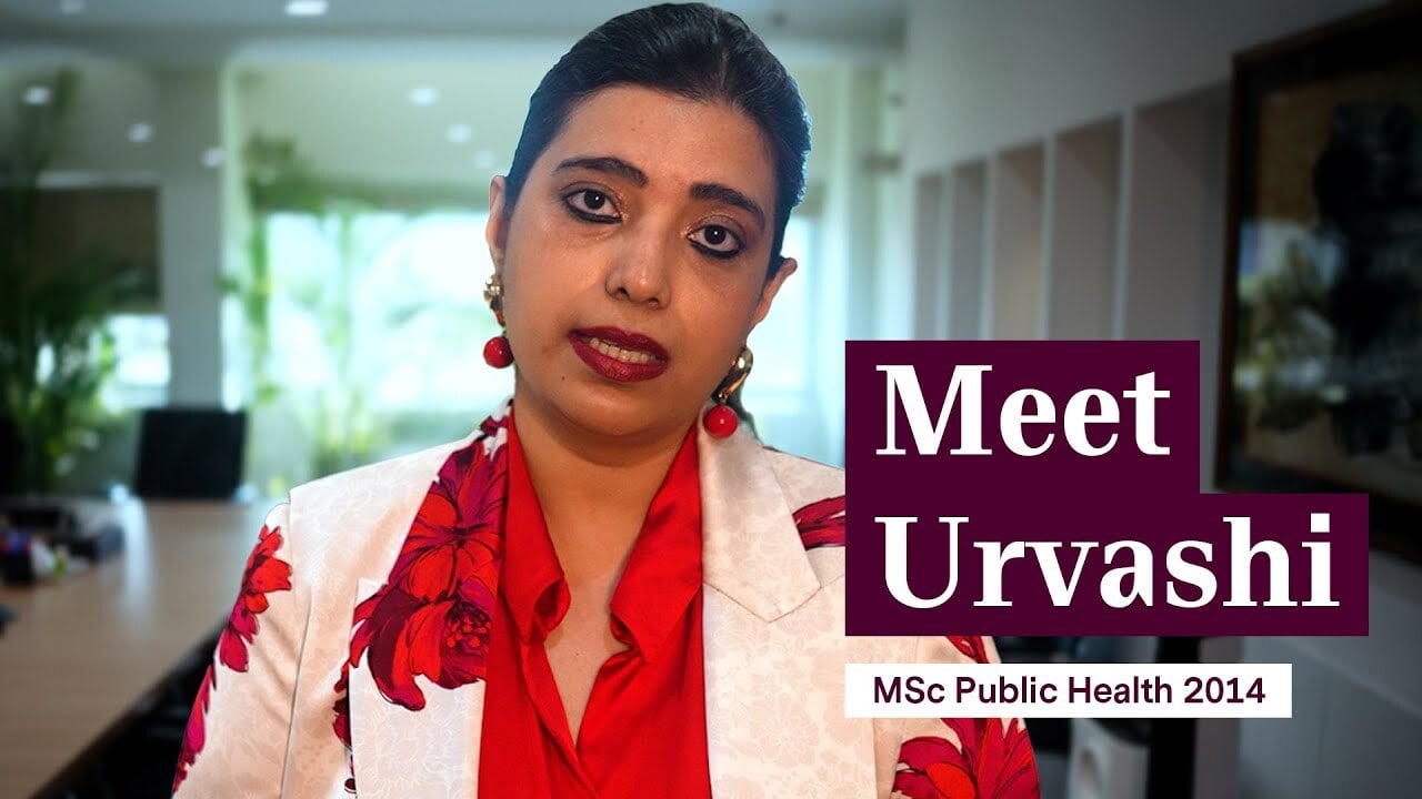Urvashi sits facing the camera wearing a red shirt and white blazer with red flowers. Text on image: Meet Urvashi (MSc Public Health, 2014)