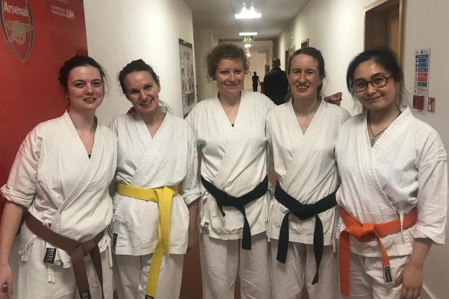 Vicky Simms is stood arm in arm with four other women in Karate uniform. Vicky and two others have black belts, one has an orange belt and the other a yellow.