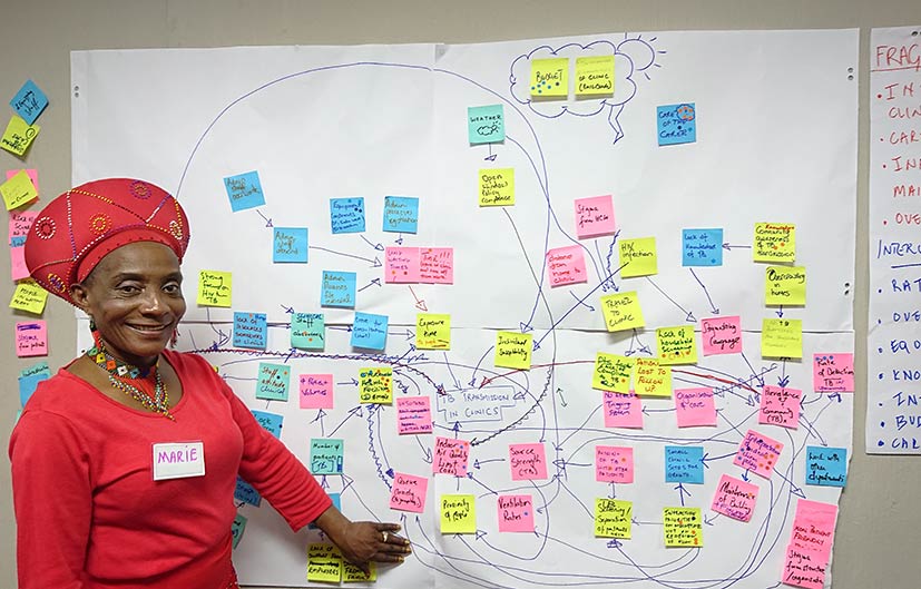 woman in red dress and headpiece standing in front of tuberculosis transmission model on chartboard paper