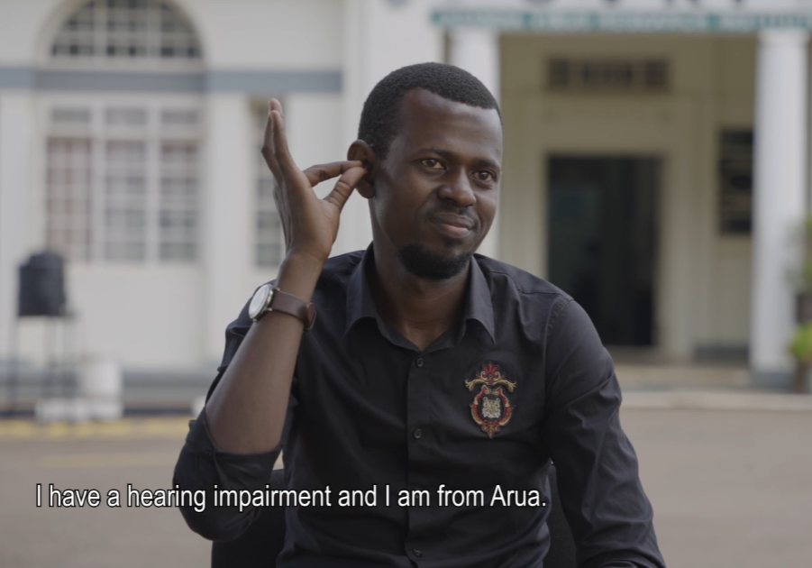 A man signs in Ugandan Sign Language. He is signing that "I have a hearing impairment and I am from Arua." He is sitting outside for a video interview.