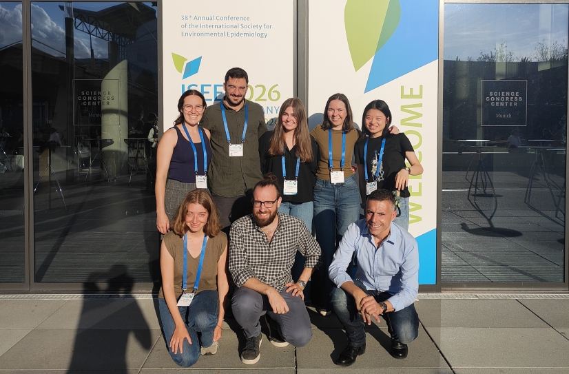 A group of 8 people, 5 standing behind and 3 kneeling in front, smiling at the camera outside a conference