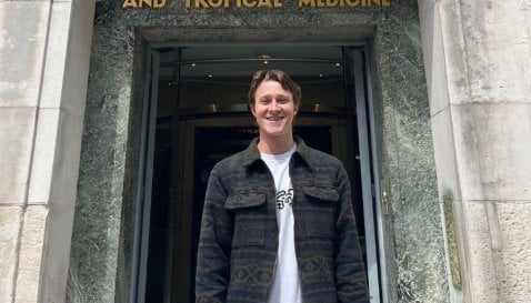 Man stands smiling outside LSHTM building with dark jacket and white t-shirt.
