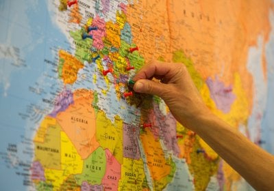 Close-up of a colorful world map focused on Europe and parts of North Africa and the Middle East, with multiple push pins marking locations. A hand is placing a green pin near southeastern Europe.