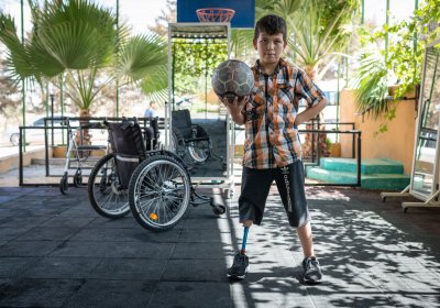 A young child with prosthetic leg standing up, holding a ball in one hand. Pictured behind is a wheelchair in the background. Photo credit: Rachel Elkind, Relief International. 