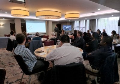 People sitting around round tables, listening to someone standing at lecturn giving presentation in hotel conference room.