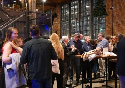 A group of people stand and talk at a networking reception inside a modern venue with exposed brick walls, high ceilings, and large industrial-style windows.