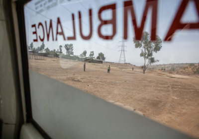 Image shows fields in Ethiopia through an ambulance window. There are people standing on the field and ambulance is written in red text across the window.