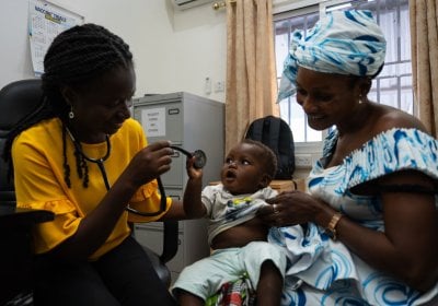 Healthcare worker using a stethoscope to examine a young child seated on a caregiver’s lap in a clinic setting,