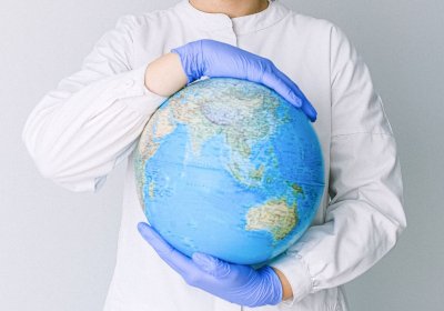 ​Photo credit: Anna Shvets, “Person With a Face Mask and Latex Gloves Holding a Globe” (Canva) 