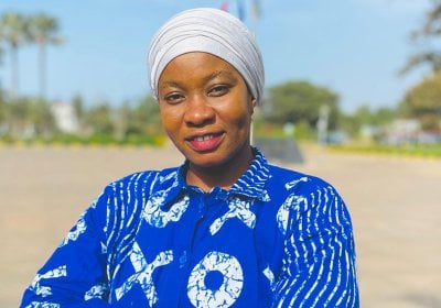 Woman with blue shirt with a white print stands arms folded with an rural landscape in the background in The Gambia.