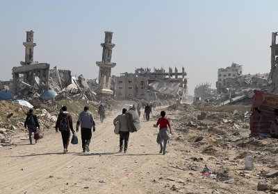 people walking along a dusty road in Gaza, carrying household items, with ruined and collapsed buildings in the background against a blue sky