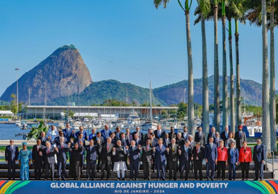 A large group of men and women in suits stand in front of palm trees, sea and mountains
