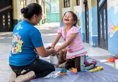 A young girl is holding hands with a woman, who is facing away towards the girl. The girl is missing her front milk teeth, which is visible as she smiles. A dog with brown fur is partially visible behind the girl. There are toys scattered around them.