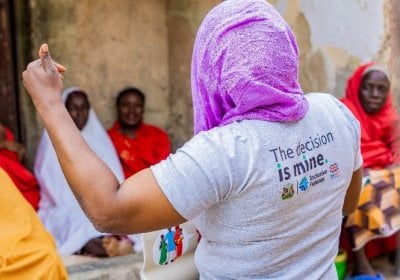 A female IFPLAN inclusive champion wearing a purple headscarf and a grey T-shirt reading ‘The decision is mine’ gestures while speaking to a small group of seated women in colourful clothing in an outdoor courtyard in Nigeria as part of an IFPLAN peer-to-peer session