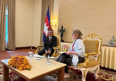 A white woman wearing a shawl and an Asian man wearing a suit sit in a formal room with a Filipino flag behind them