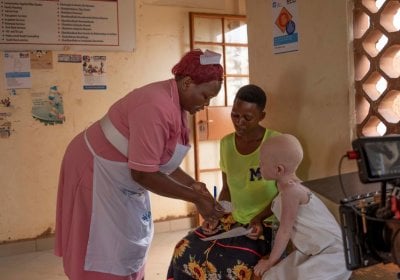 A nurse at a health facility in Luuka district, Uganda is assisting a mother of a child with albinism. She is handing over a prescription to the mother and her daughter, who is sitting next to her looks at the piece of paper. In the corner, a film camera is visible.