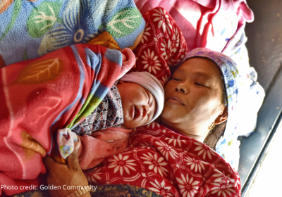 A nepalese mother is laying with her newborn on a bed surrounded by brightly coloured sheets.