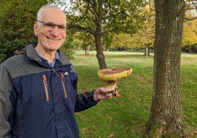 Image of a man standing in a park with trees and grass. He is wearing a blue and grey jack, and glasses, and holding a large mushroom in his hand outstretched.