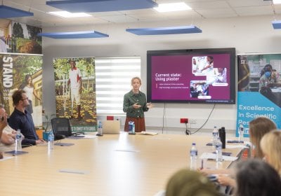 A presenter speaks during the STAND project inception meeting at MRC Gambia, showing a slide on plaster use while attendees listen around a conference table. The STAND initiative supports amputees in The Gambia.