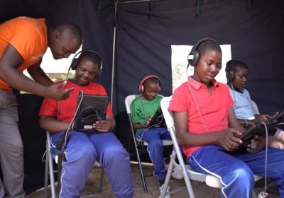 Four adolescents sit with electronic tablets and headphones completing questionnaires. A man stands beside one of the boys, supporting them on the tablet.