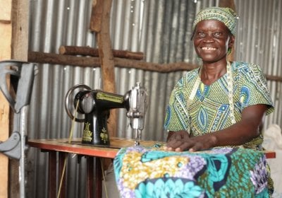 Mary is a refugee from South Sudan living in Kenya. She has a physical disability and settled in Kalobeyei where she has a business. In the photo, she is sewing kitenge print clothes behind a sewing machine inside a room. Her walking stick is placed against the wall, which is made of metal sheets and wood.