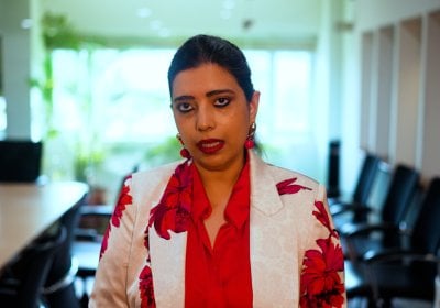 Urvashi sitting in a meeting room wearing a red shirt and white blazer with red flowers.