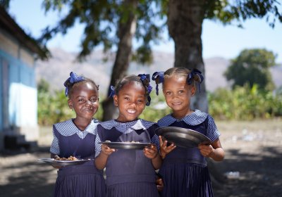 Three young girls in school uniform holding plates of food. Photo: WFP/Antoine Vallas