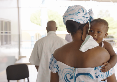 A mother walks away from camera holding her baby who looks towards the camera
