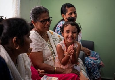 A child smiles towards the camera while sitting on a woman's knee. Another woman sits behind her while a different woman sits in front of her